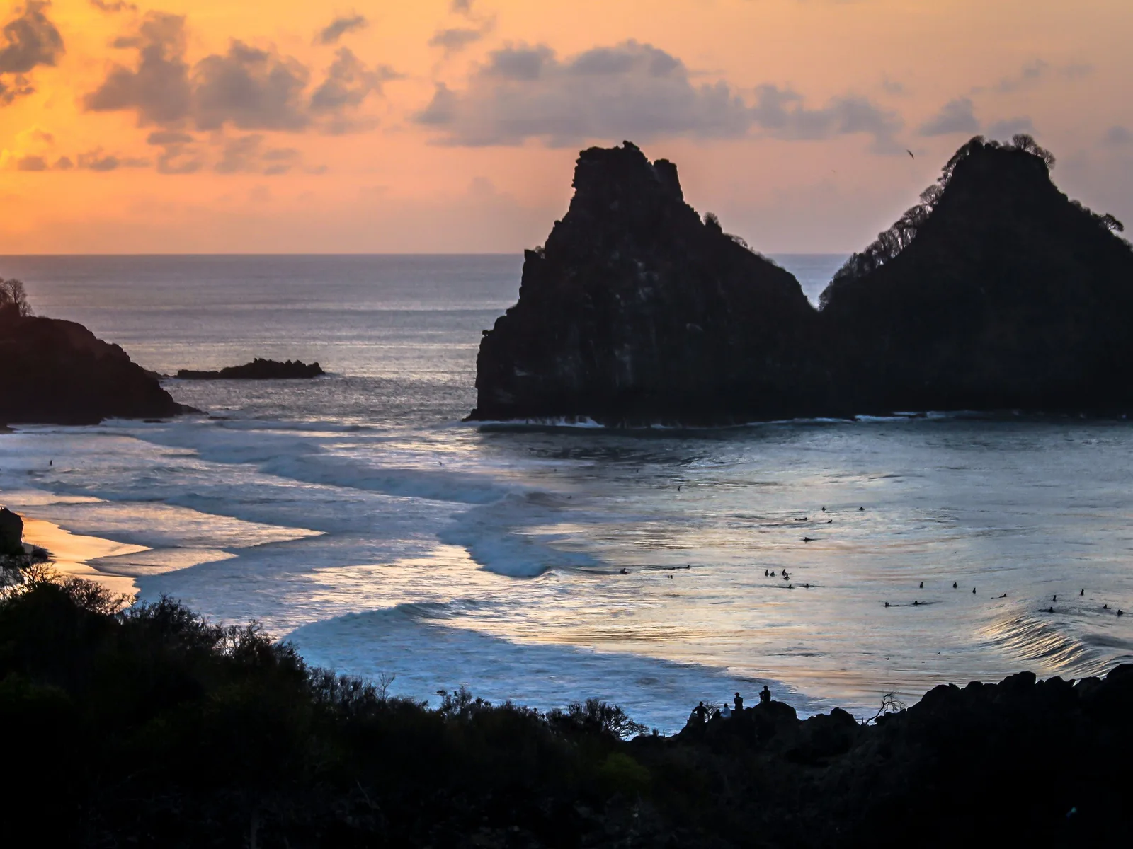 Morro Dois Irmãos in silhouette against an orange sunset at Cacimba do Padre, with surfers sitting out in the water and wet sand reflecting the sky