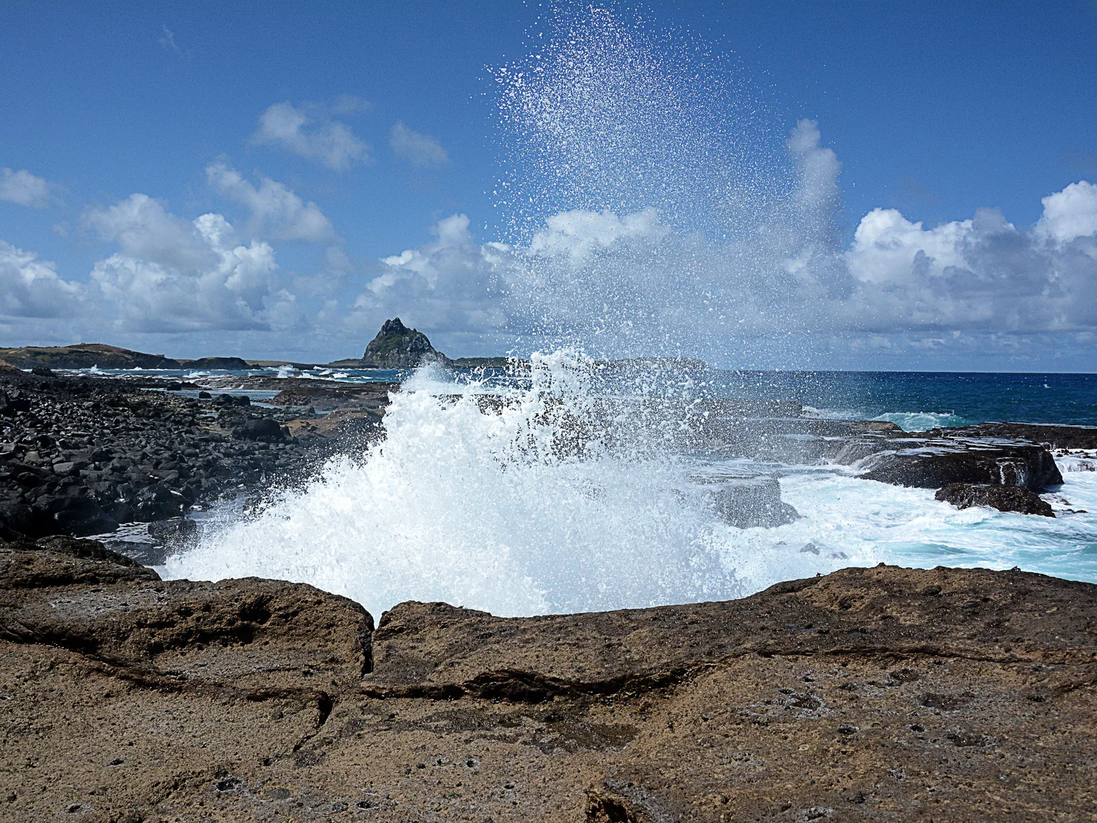 A wave bursting white and high against black volcanic rock at the shoreline, spray thrown up against a blue sky