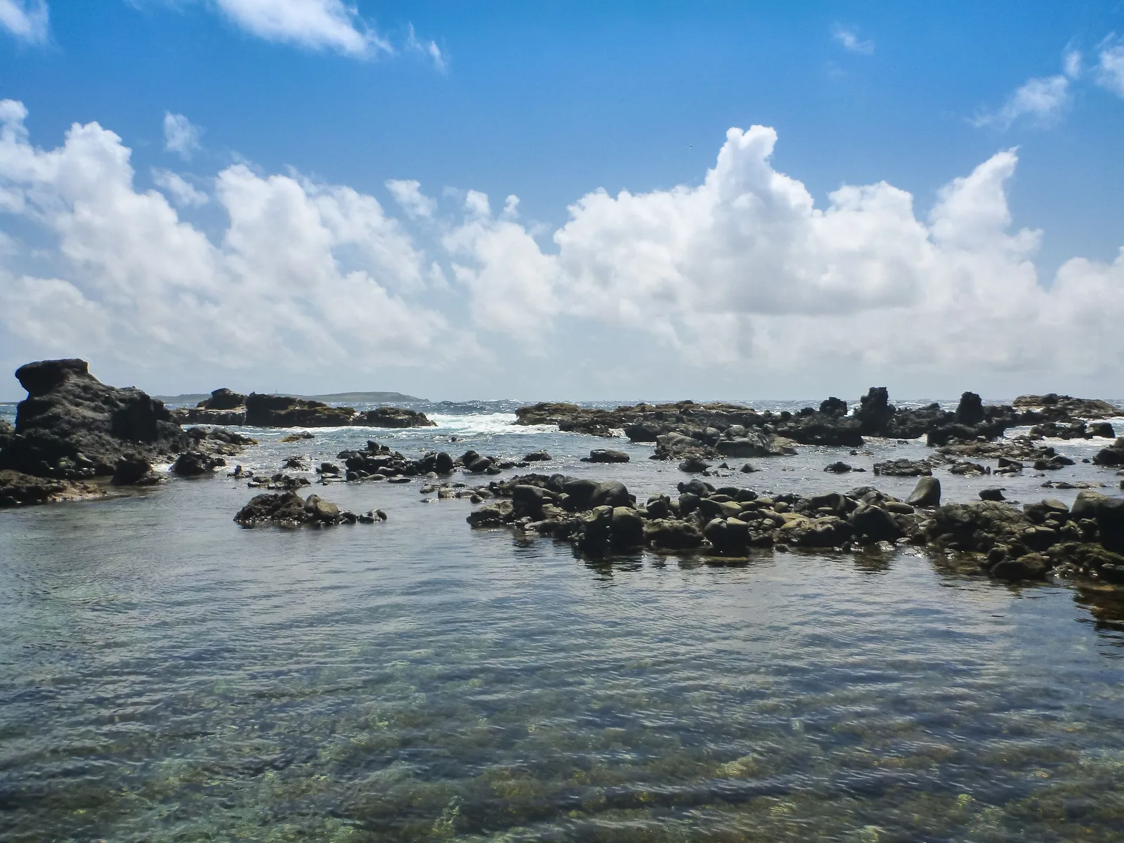 A broad shallow reef flat of clear water with dark volcanic rock breaking the surface across it, under a sky of tall white cumulus