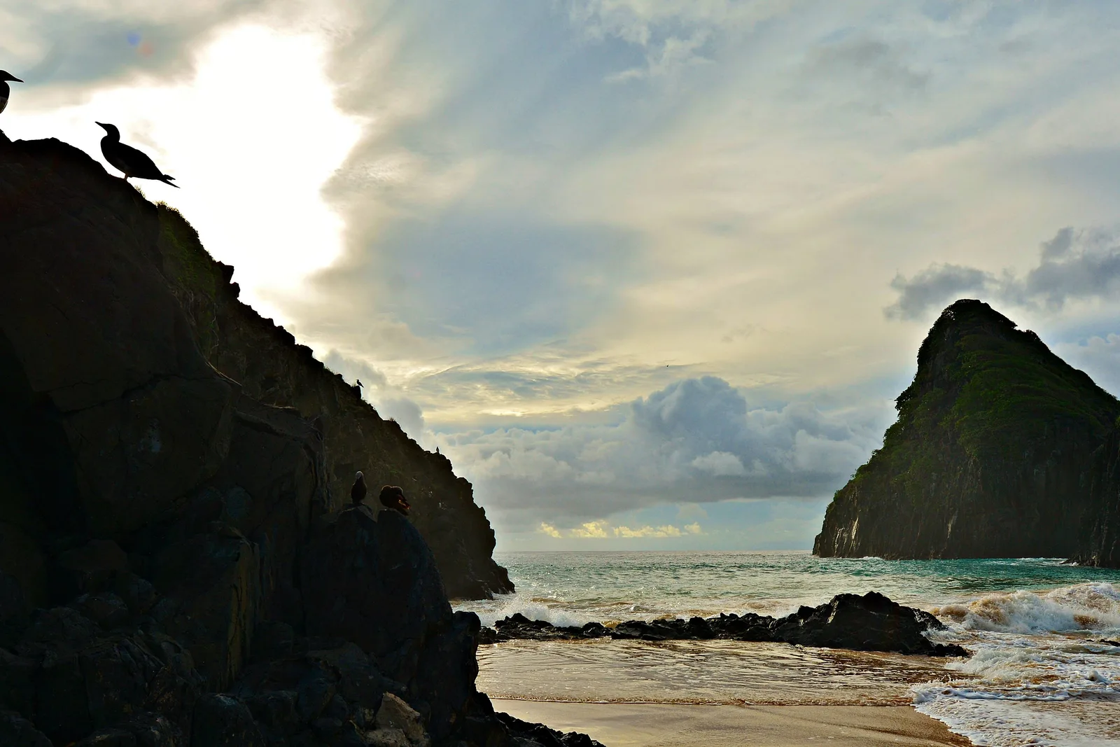Birds on a dark rock in silhouette at Cacimba do Padre, two on the skyline and two lower down with two people sitting beside them, and a single peak of Morro Dois Irmãos on the right against a bank of backlit cloud
