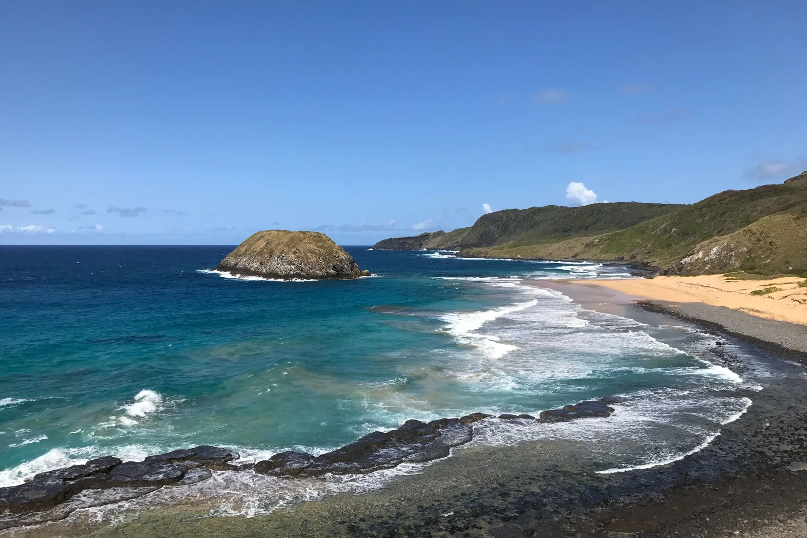 Praia do Leão in the morning: a dark rocky island standing off a long curve of pale sand, with surf breaking over a shallow reef in front of it