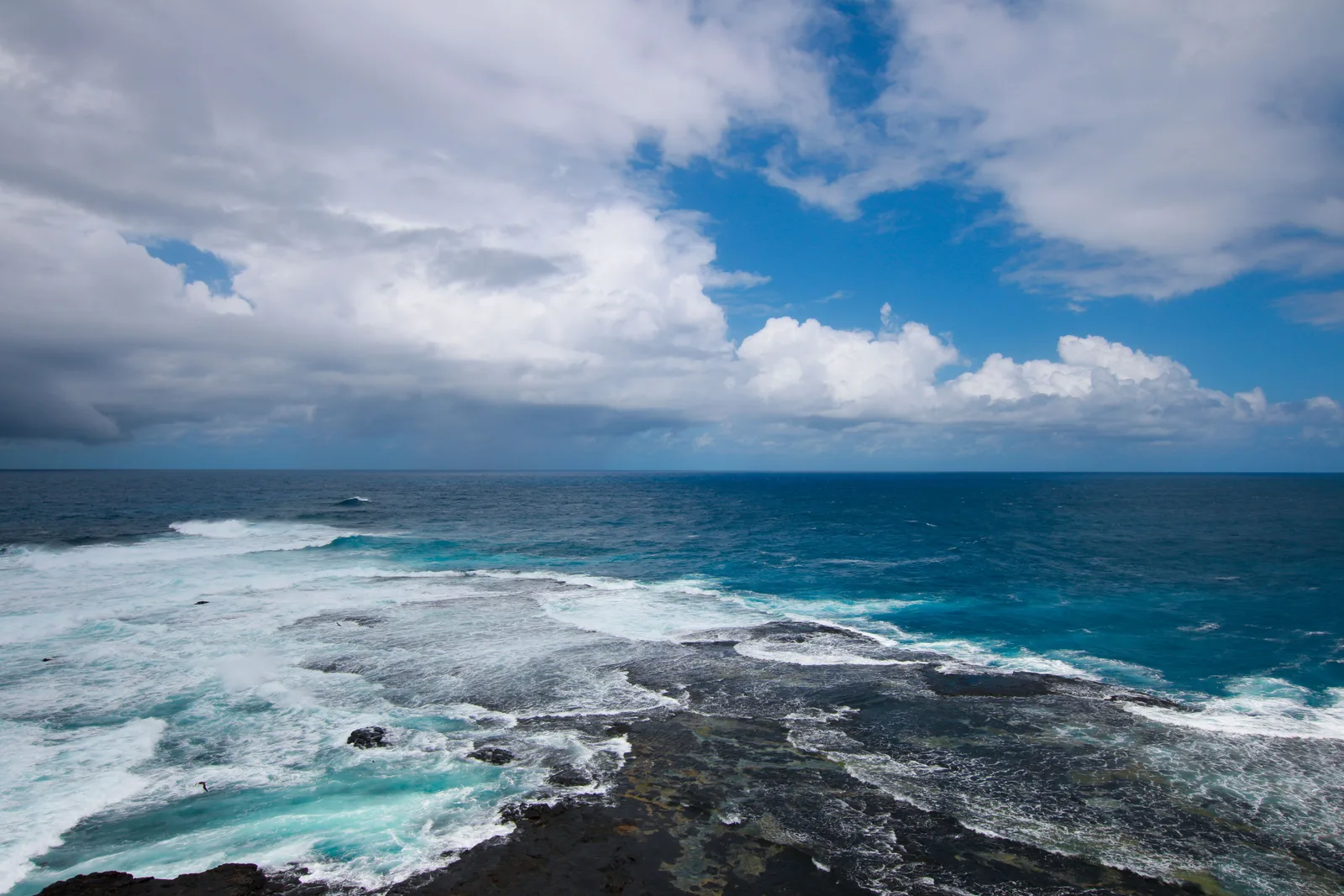 White surf breaking over black volcanic rock at Ponta das Caracas under a sky of scattered cloud, with open ocean running to the horizon