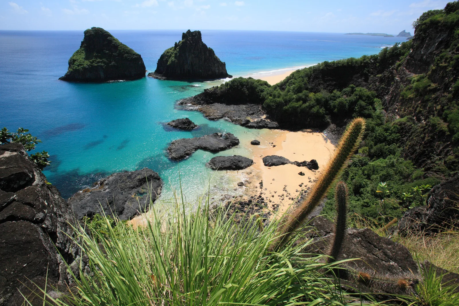 Baía dos Porcos from the clifftop path: coarse grass in the foreground, black volcanic rock, a small strip of sand and the twin peaks of Morro Dois Irmãos standing in blue-green water