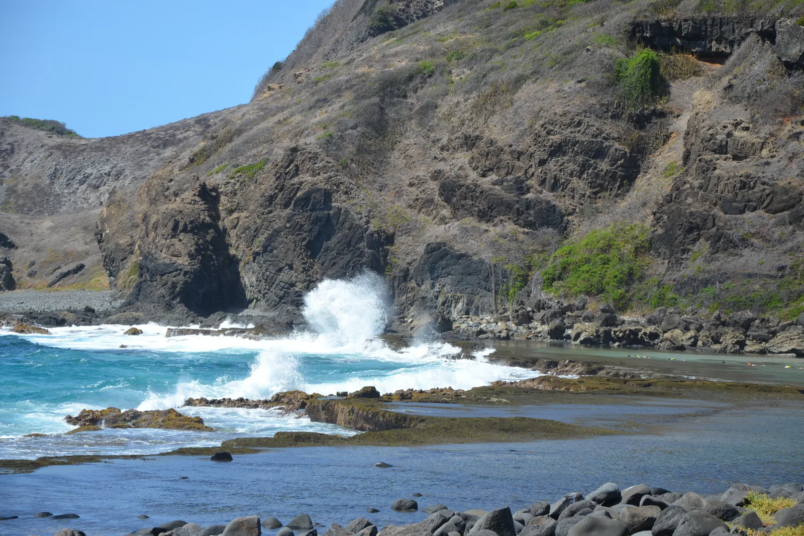 A wave bursting white against dark rocks at the foot of a steep brown-gray headland with sparse scrub on it, a shallow rock pool in the foreground
