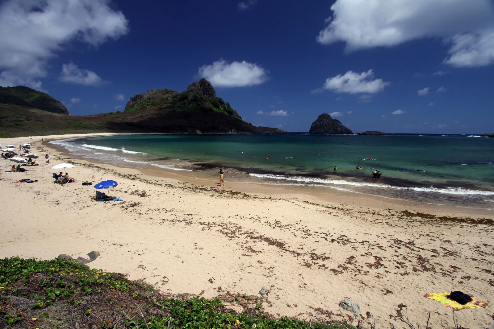 A wide sandy beach at Baía do Sueste with a blue parasol and a few bathers, a rounded green hill behind and a pointed rock standing offshore