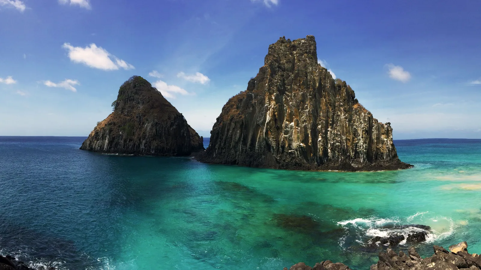 The twin volcanic peaks of Morro Dois Irmãos rising straight out of clear turquoise water at Baía dos Porcos, with dark rock breaking the surface in the foreground and open blue sea behind