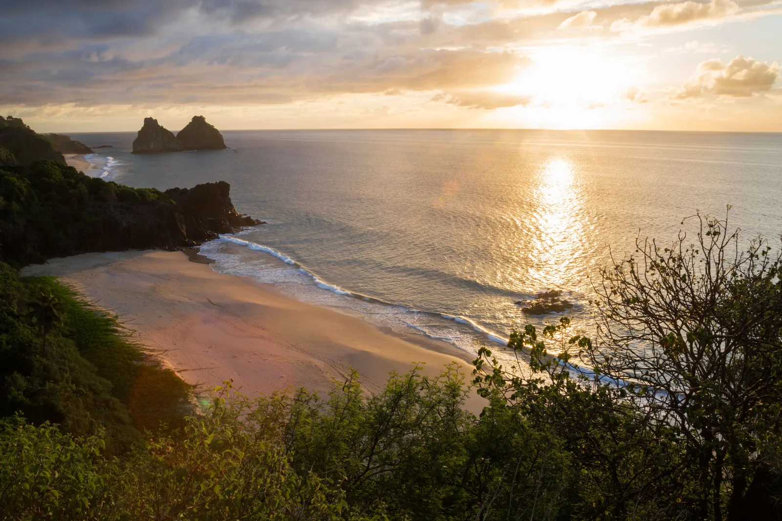 The sun low over the sea seen from a clifftop above Praia do Boldró, warm light across the water and the beach curving away below