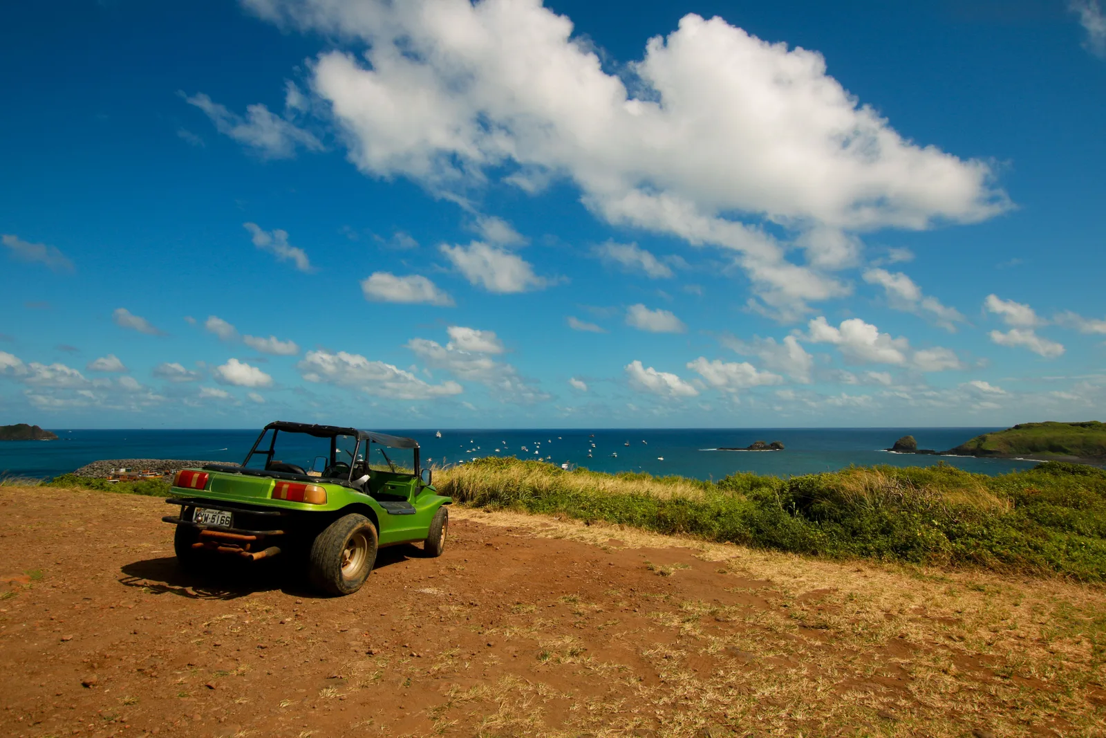 A green open-topped buggy parked on a dirt track on a headland above the sea, under a blue sky with white cumulus