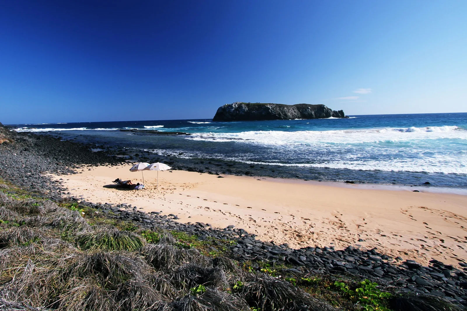 Praia do Leão from the rocks at one end, a long pale beach with a dark island standing offshore and pale green water over the reef