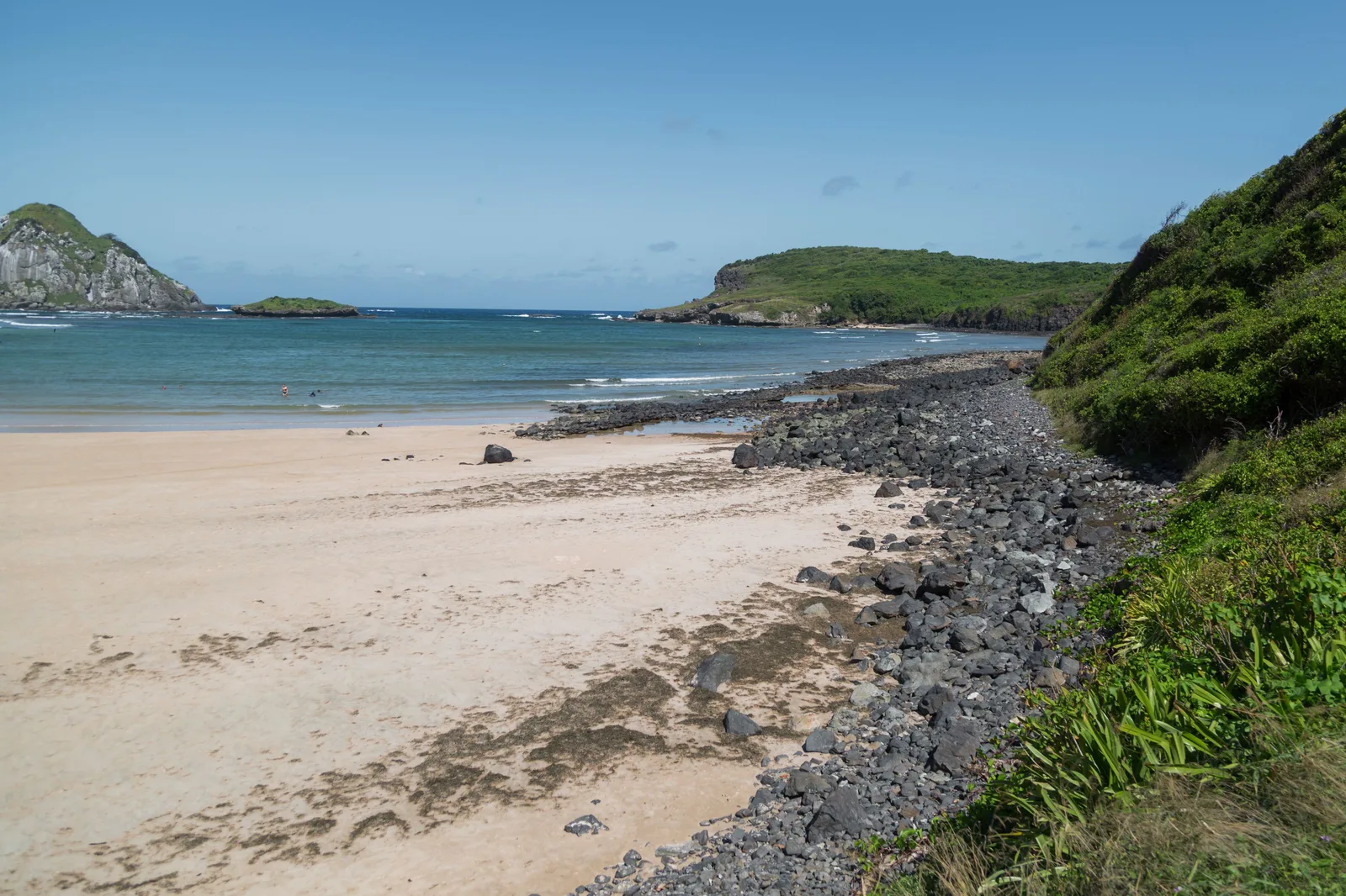 The beach at Baía do Sueste: pale sand and scattered dark rocks in the foreground, a green hill on the right and low islands out on the water