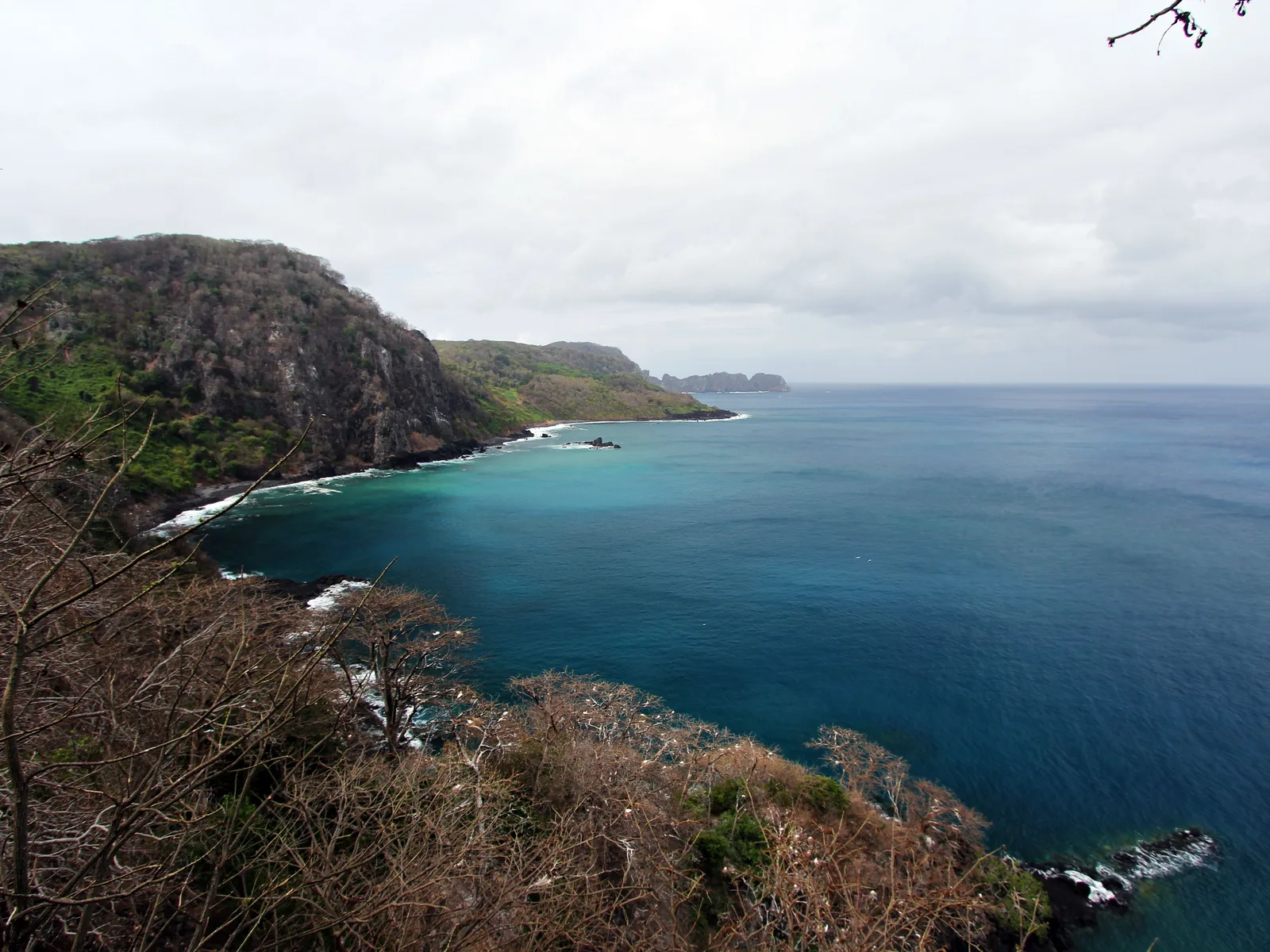 Baía dos Golfinhos seen from high on the cliff above it: an enclosed blue bay with a wooded headland running out around it and no way down to the water