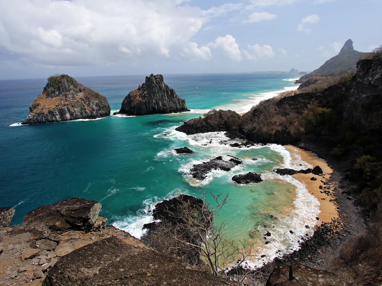 Baía dos Porcos from above: the twin peaks of Morro Dois Irmãos on the left, a small strip of sand, black volcanic rock breaking through pale green shallows and white surf beyond