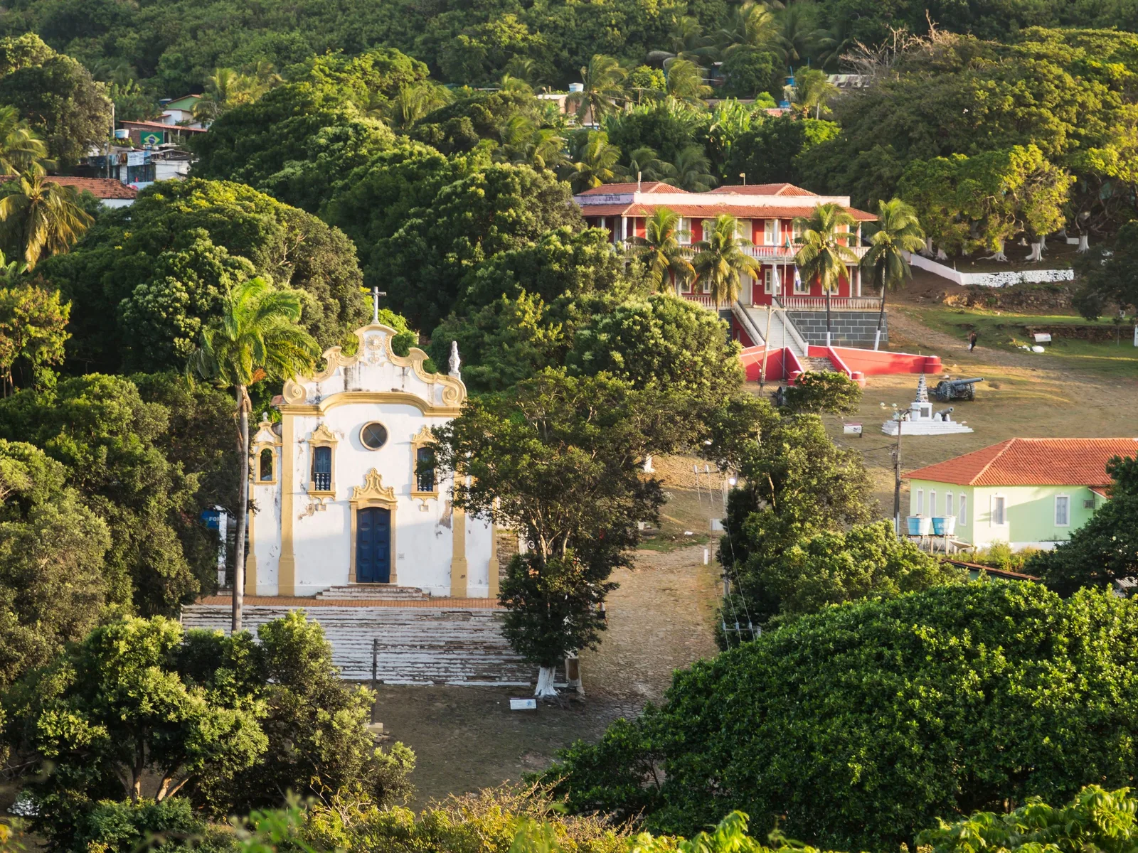 The white and yellow church of Nossa Senhora dos Remédios seen from above among tall trees and palms, its door blue, with the red and cream buildings of the village around it