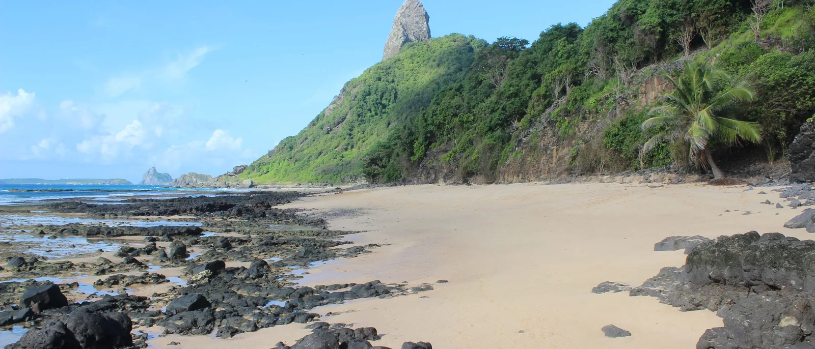 The beach at Praia do Boldró in a wide frame: pale sand and black volcanic rock in the foreground, a steep green hillside and the peak of Morro do Pico rising behind it