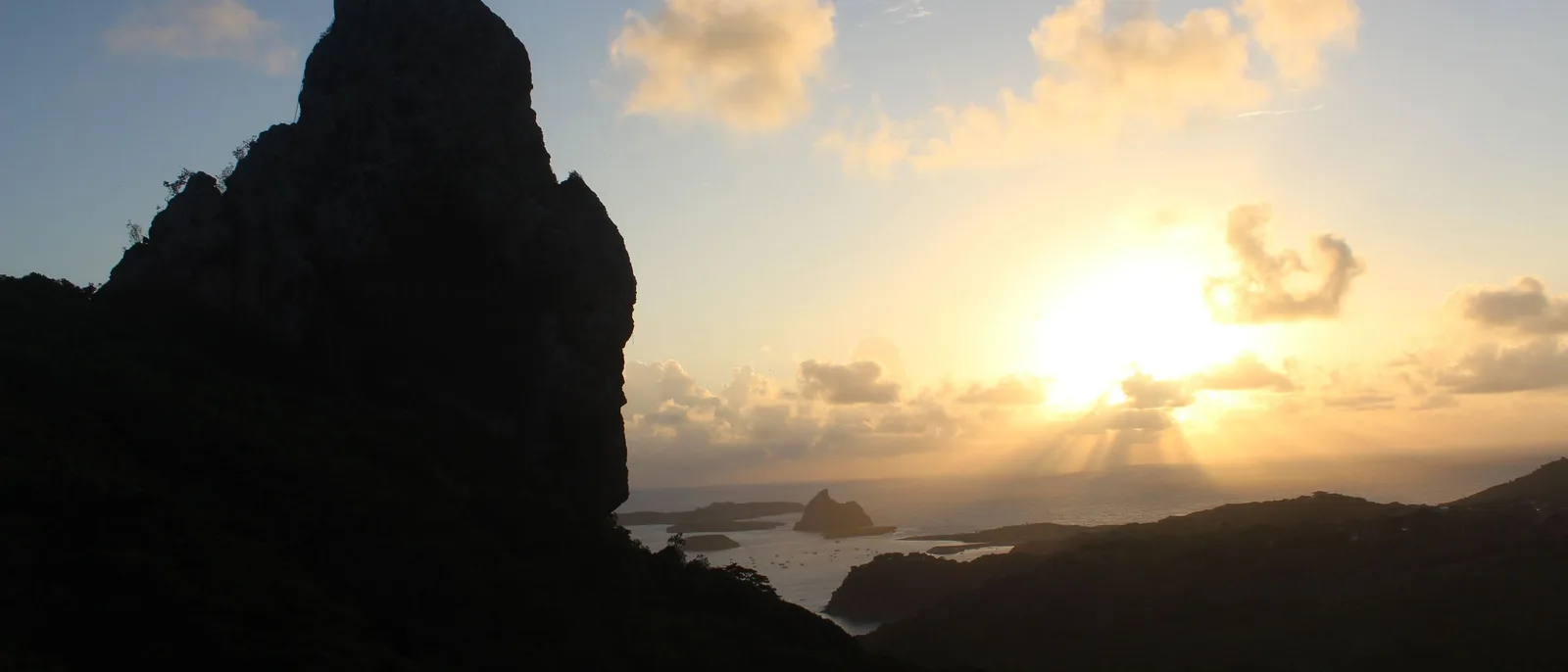 The dark pointed silhouette of Morro do Pico against a low sun and broken cloud, seen across the wooded interior of the island with the sea beyond