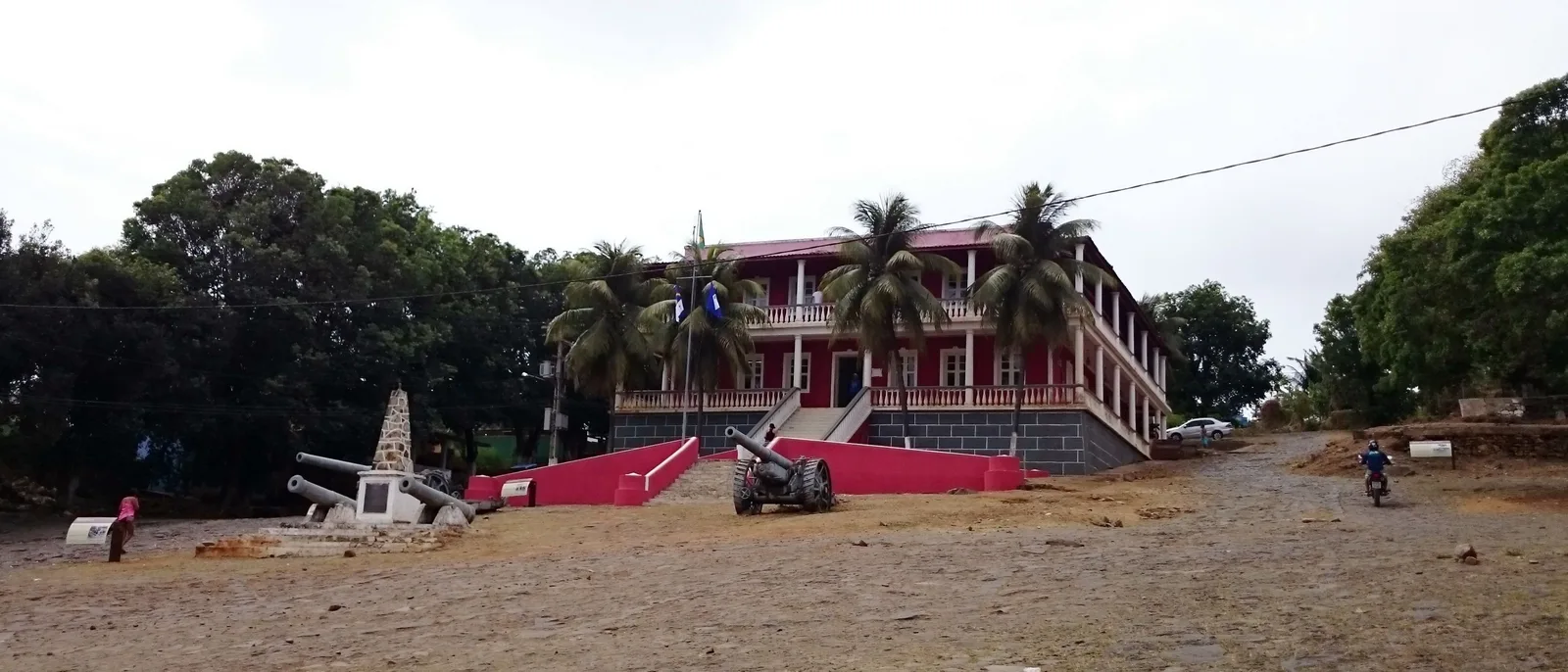 The red and white two-story administration building of Fernando de Noronha behind a row of palms, with old cannon and a monument on the red-earth plaza in front of it