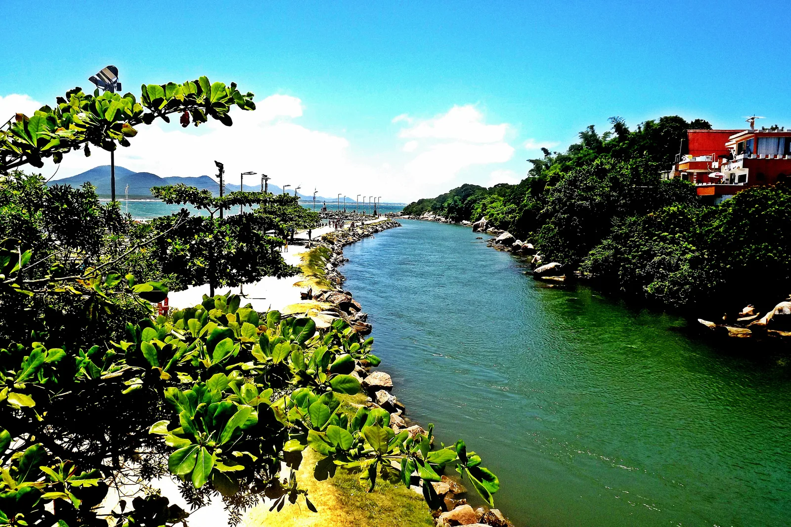 The green channel at Barra da Lagoa winding out to the open sea past a stone walkway, lush vegetation framing the view and mountains beyond