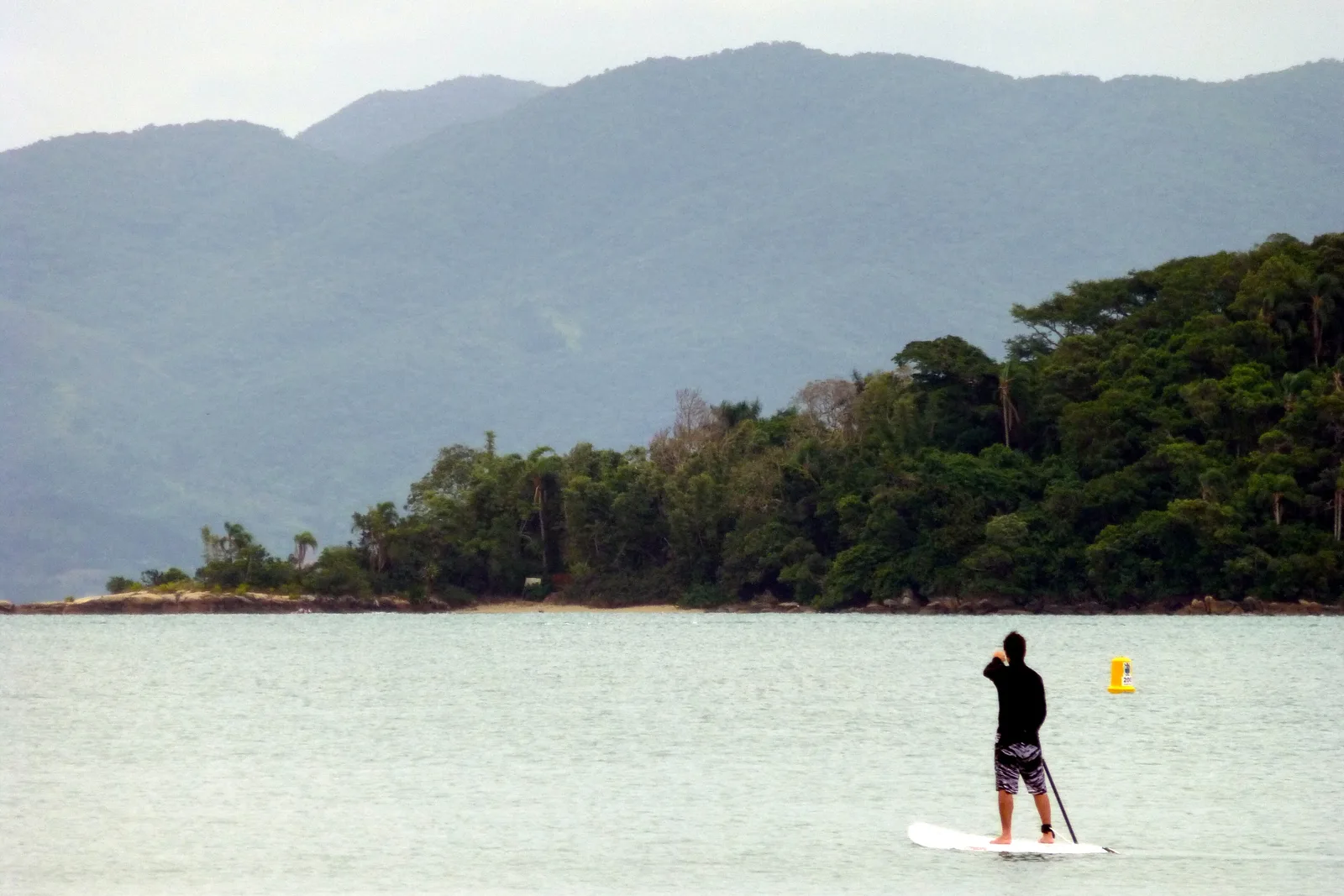 A stand-up paddleboarder on flat, still water with a small forested island and green hills rising behind