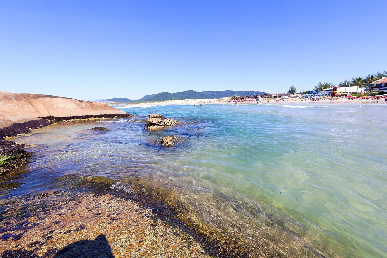 Clear shallow water over dark rocks at the end of Praia da Joaquina, the beach and the hills rising behind it