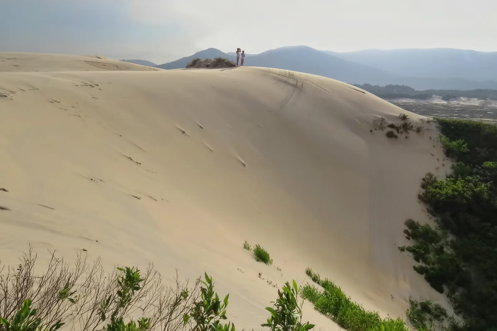 Two people standing on the crest of a tall sand dune above the Lagoa da Conceição, hills hazy in the distance