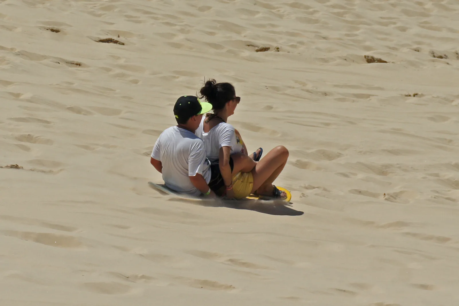 Two people sliding down the tall dunes behind Joaquina on a sandboard