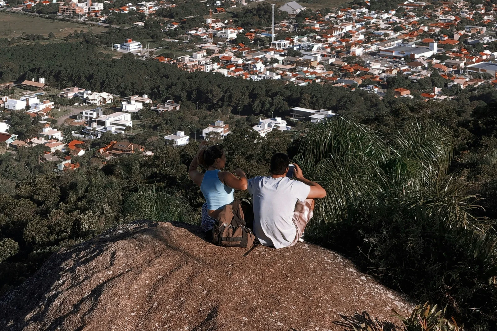 Two hikers sitting on the summit rock of Morro do Lampião, photographing the Campeche neighborhood spread out in the forest below