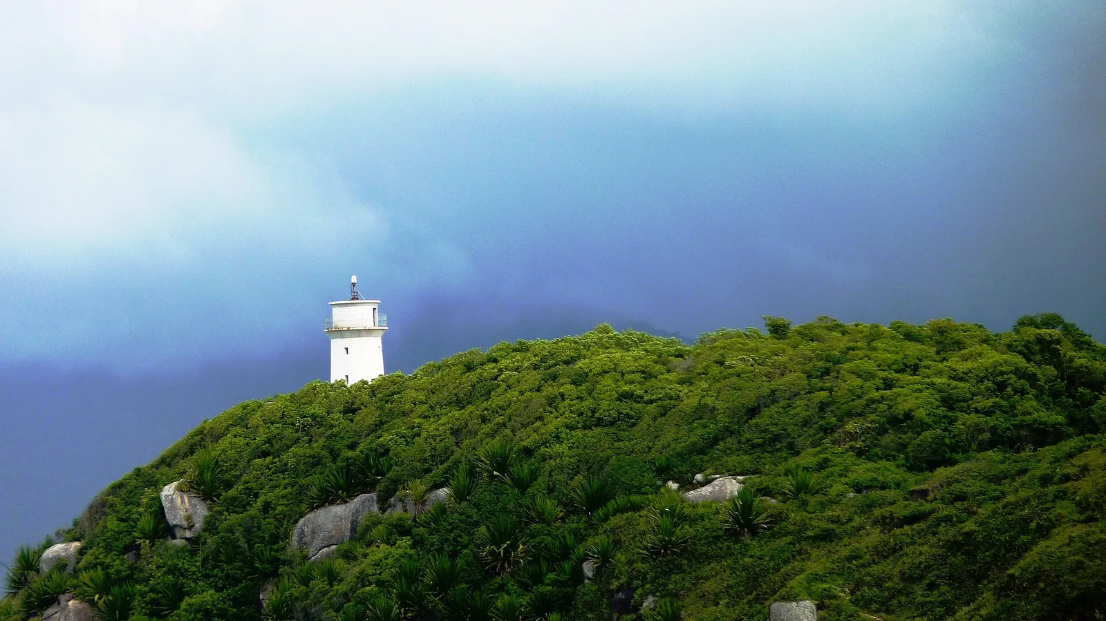 The white Naufragados lighthouse on a green headland above the beach, storm clouds building over the strait