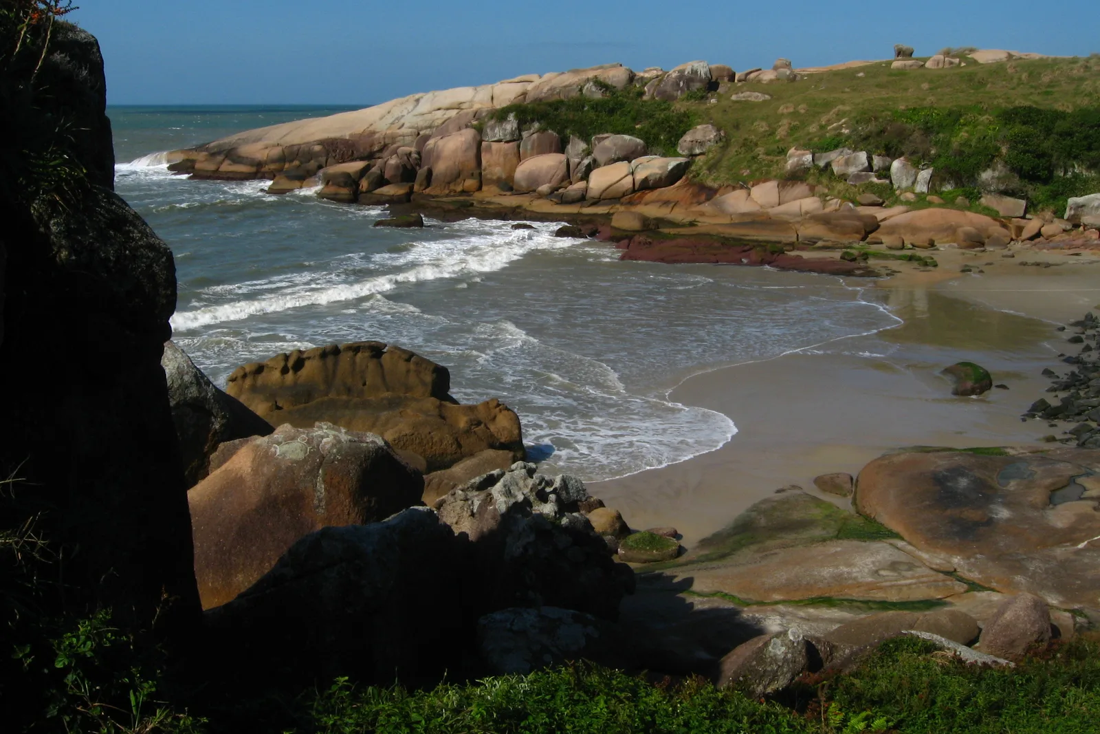 The small enclosed cove of Praia da Gravatá — a strip of sand ringed by pink granite boulders and a low green headland