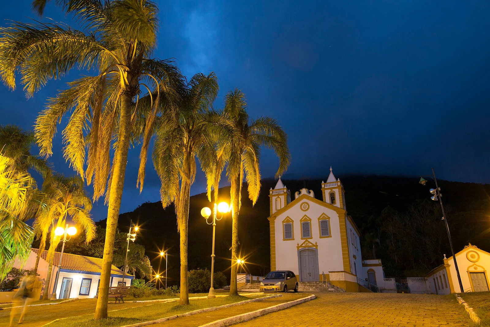 The lit white colonial church at Ribeirão da Ilha against a deep blue dusk sky, tall palms around the square