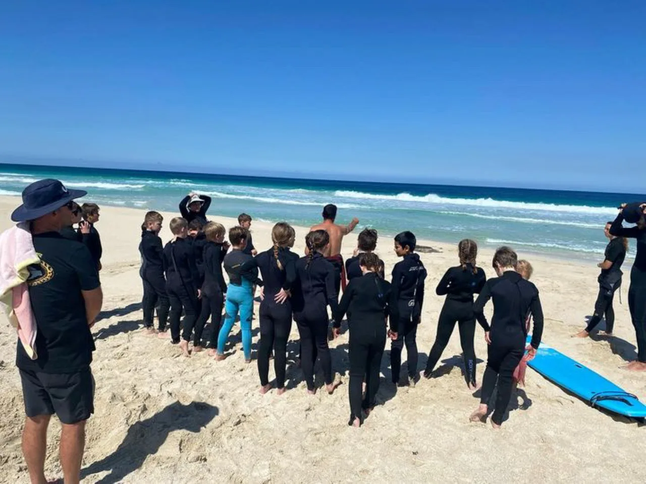 A group of students in wetsuits standing on the sand around an instructor, boards down, listening to the briefing before a surf lesson