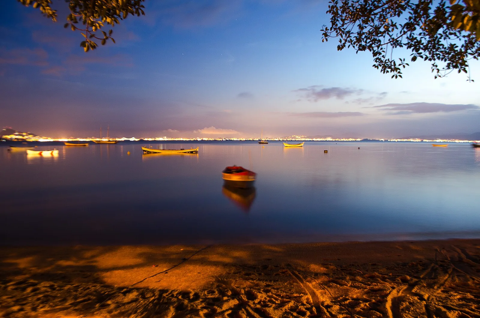 Sunset over flat water at Praia da Tapera, small boats silhouetted and the shore lights coming on across the bay