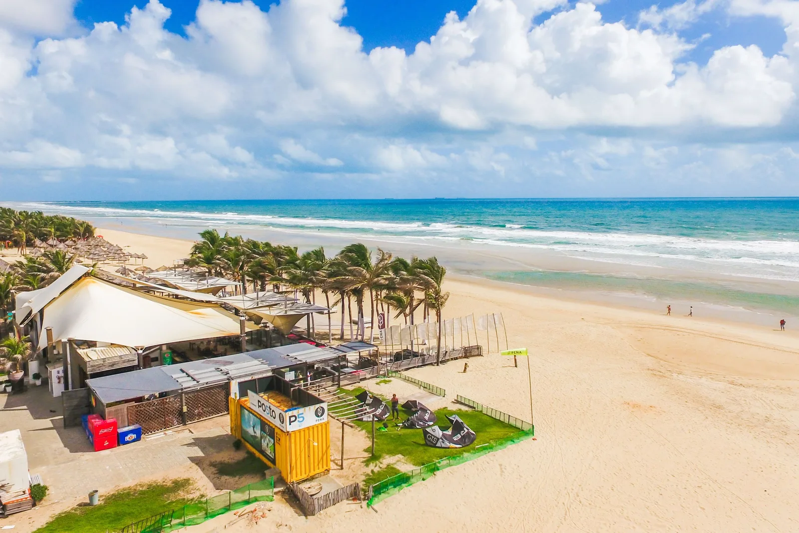 A beach barraca at Praia do Futuro seen from the sand: a white canopy and a low building among coconut palms at the top of the beach, with a wide empty stretch of sand and green surf beyond