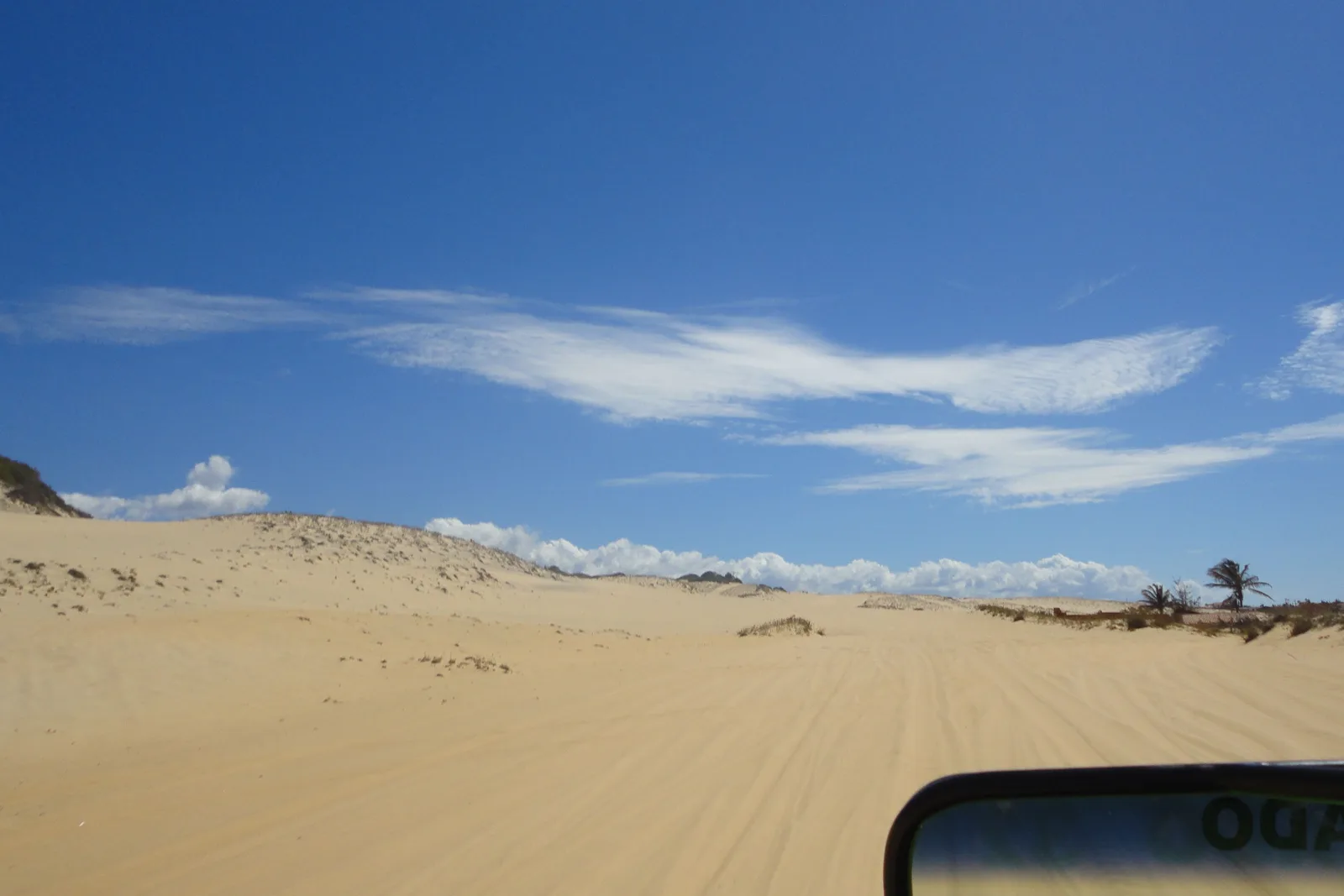 A wide pale dune field seen from inside a beach buggy, with the edge of the vehicle in the bottom corner of the frame and low green scrub along the far side of the sand