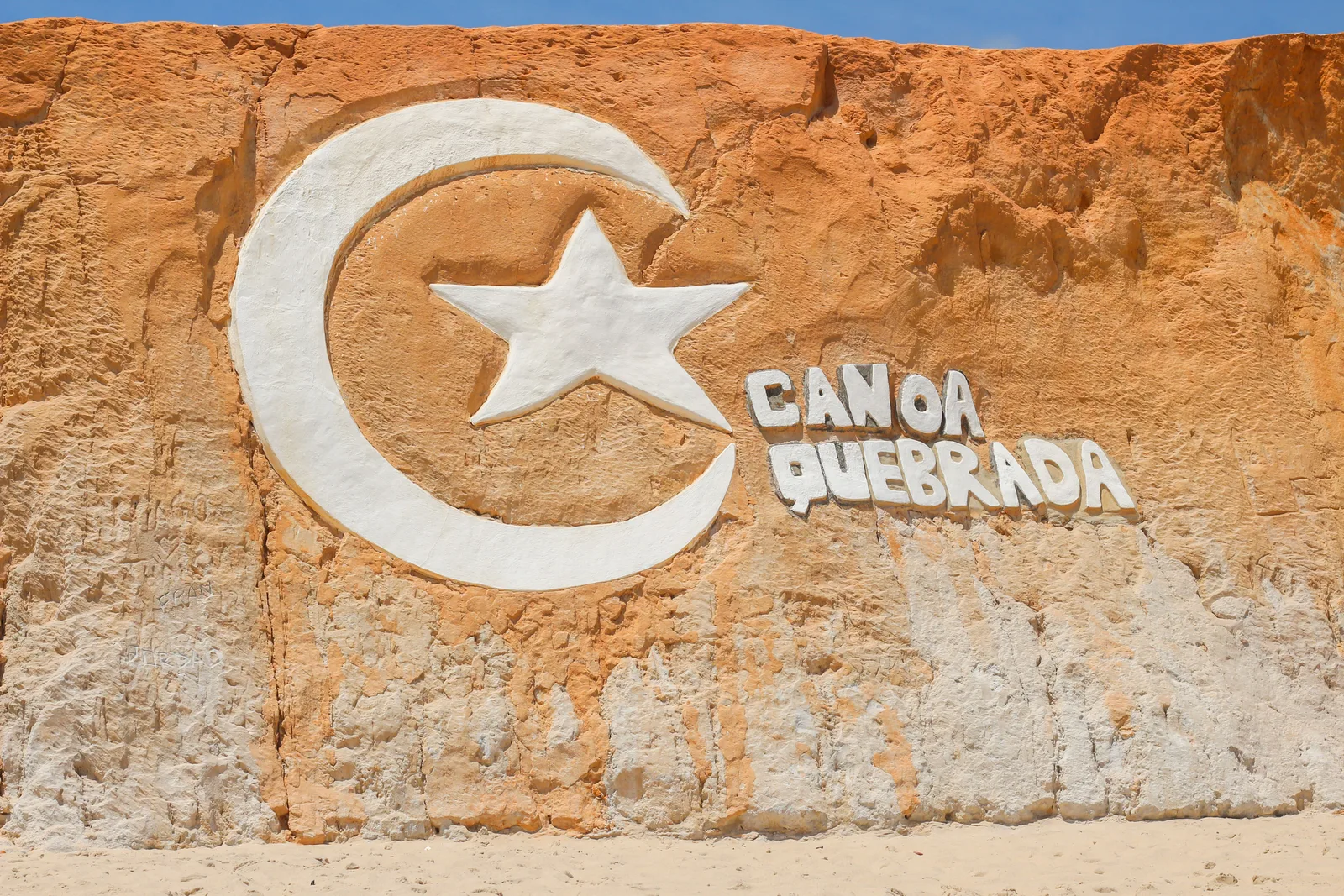 A crescent moon and star carved in relief into the face of a red and cream sandstone cliff at Canoa Quebrada, with the words Canoa Quebrada cut beside them