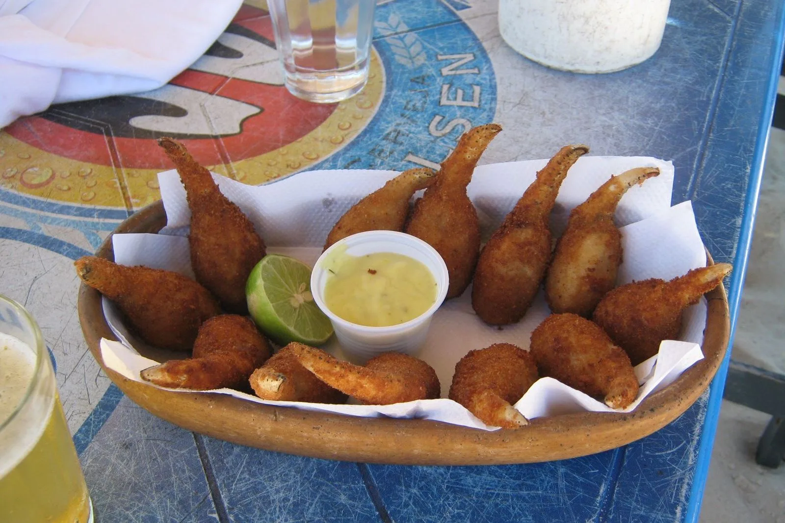A wooden bowl of breaded fried crab claws with the pale claw tips still showing, a half lime and a pot of pale sauce in the middle, on a blue beach table with two glasses of beer beside it