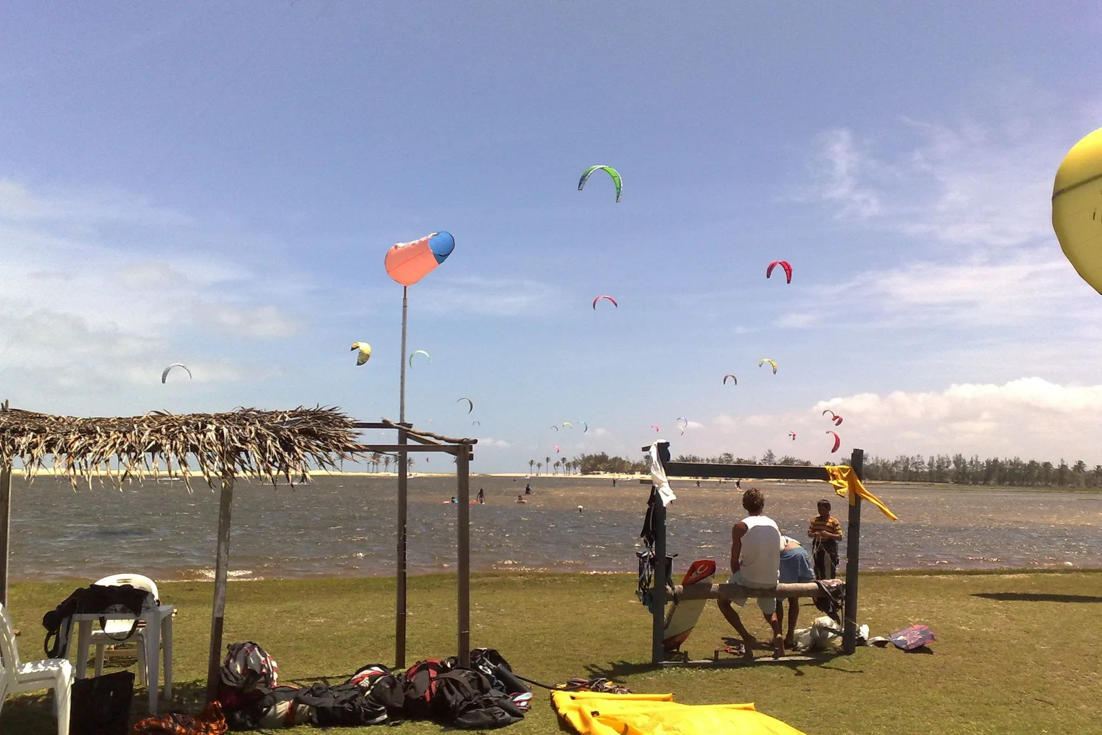 Kitesurfing kites in the air over the Lagoa do Cauípe with more kites laid out on the grass beside a thatched shelter, and two people rigging a kite in the foreground