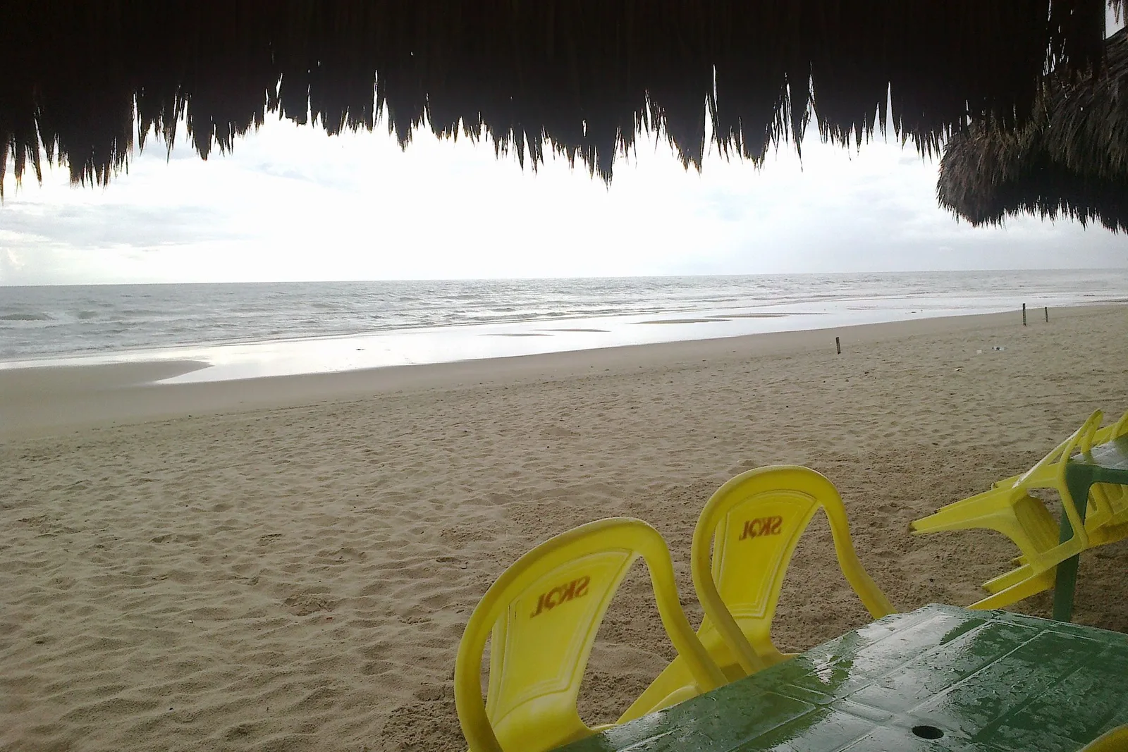 The view out from under a palm-thatch roof at a Praia do Futuro barraca: the fringed edge of the thatch across the top of the frame, yellow plastic chairs on the sand and a wide flat beach beyond