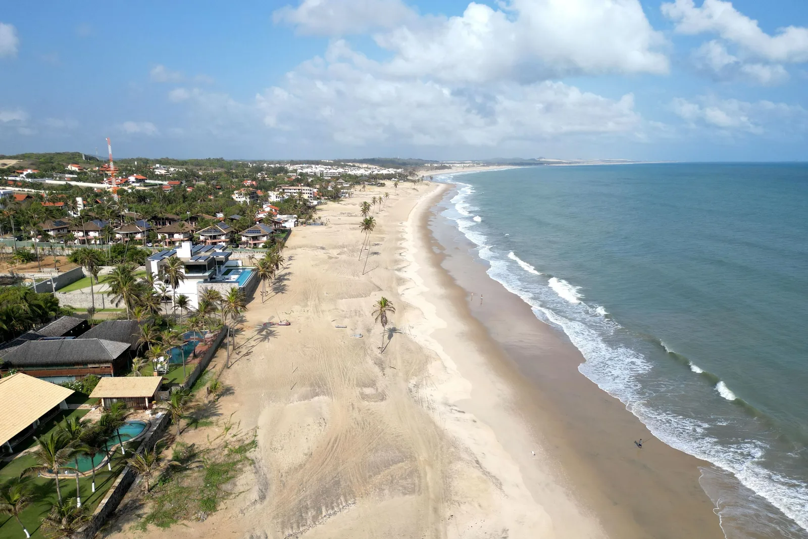 Cumbuco from the air: a long curve of pale beach with surf breaking along it, the village of low buildings and palms set back behind the sand, and dunes beyond