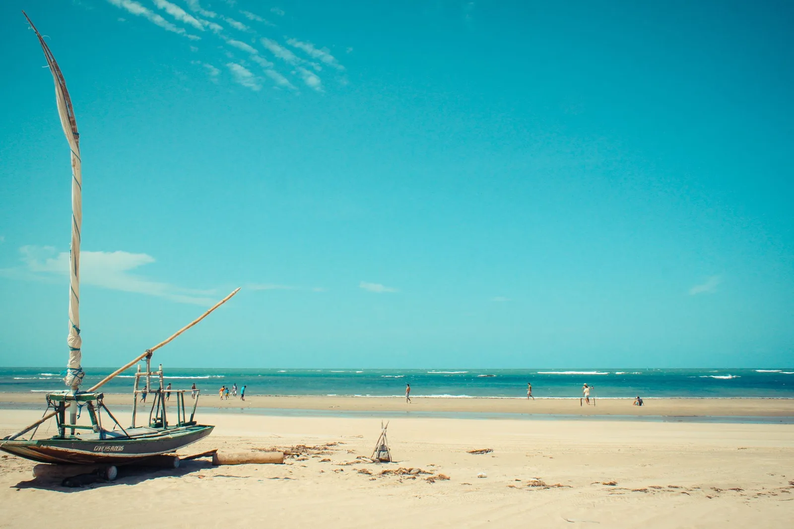 A jangada resting on wide white sand at Flecheiras with its mast bare, under a big blue sky with pale turquoise water running out to the horizon