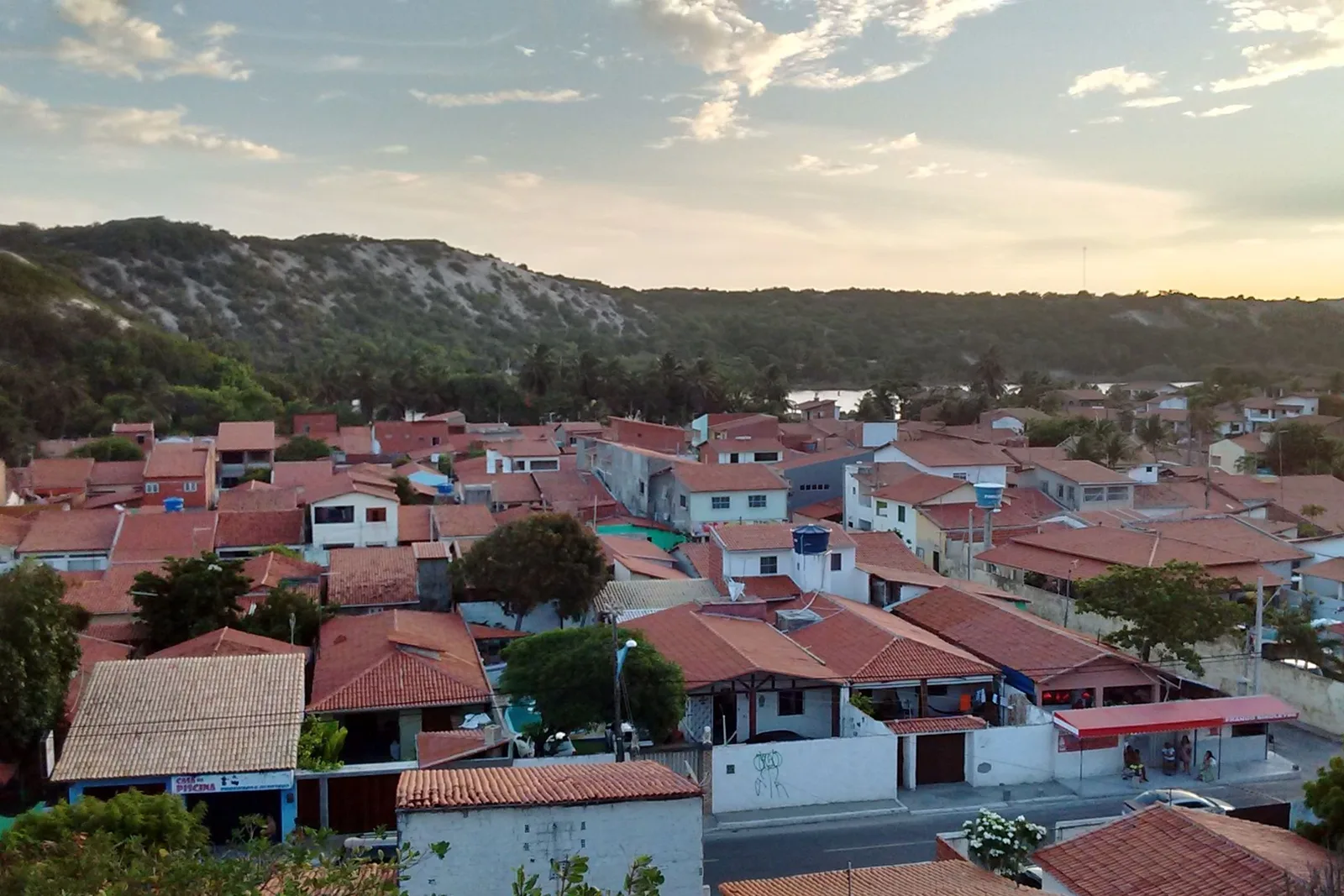 The village at Praia do Iguape from above: rows of red-tiled roofs among trees with a pale dune ridge rising directly behind the houses