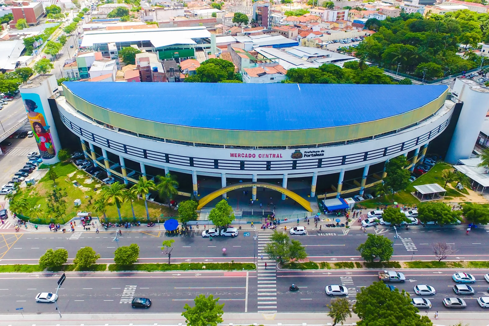 The Mercado Central de Fortaleza from the air: a long curved building with a bright blue roof and a yellow and blue front, standing beside a wide avenue with the city behind it