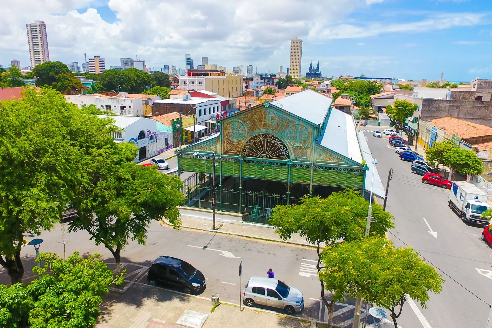 The Mercado dos Pinhões from above: a low green cast-iron and glass pavilion with an ornate gable standing on a corner among trees, with the old center and the cathedral spires behind it