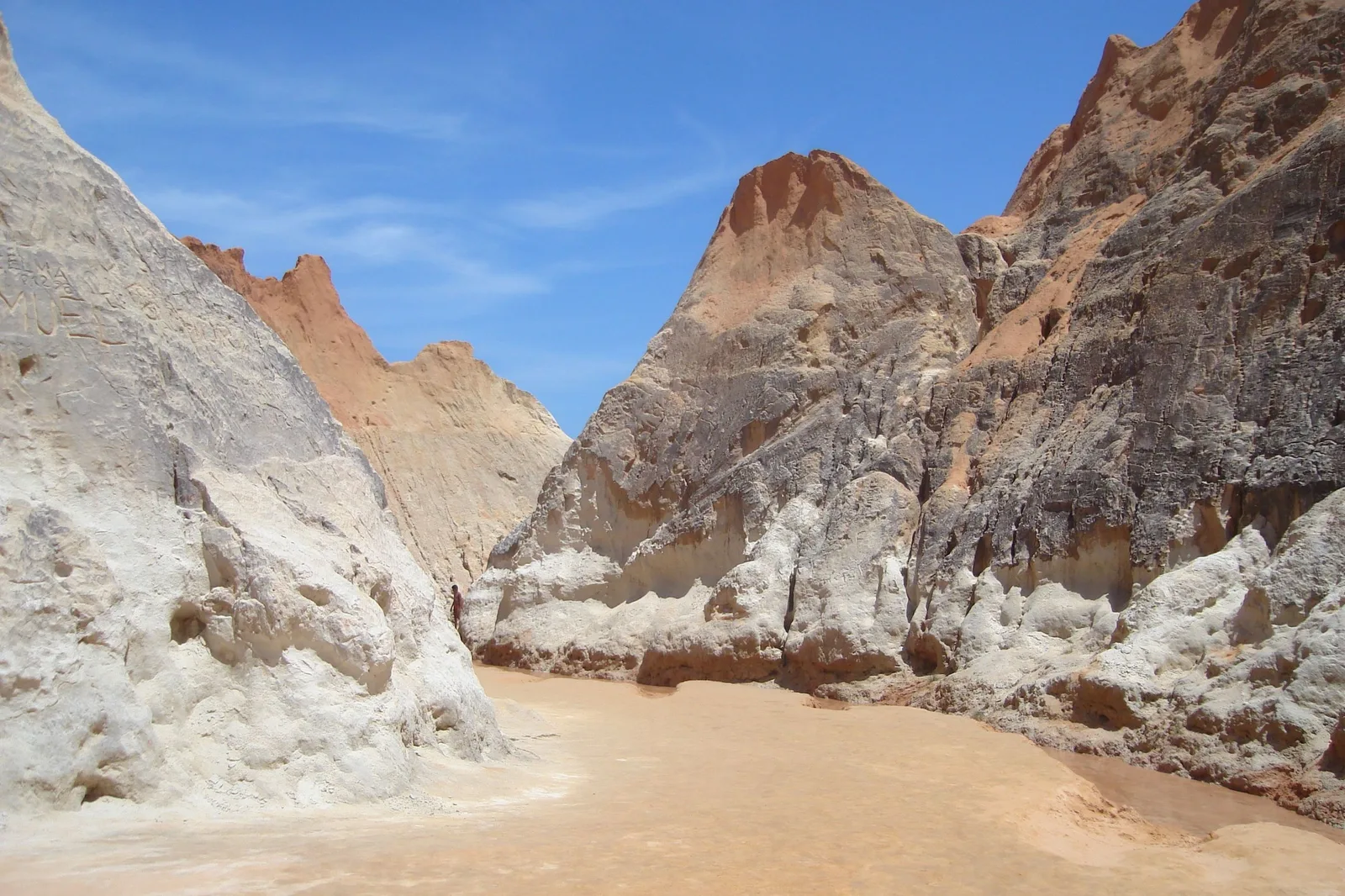 A narrow passage between tall pale sandstone cliff walls at Beberibe opening onto a floor of dry sand, the rock streaked white, cream and pink under a blue sky