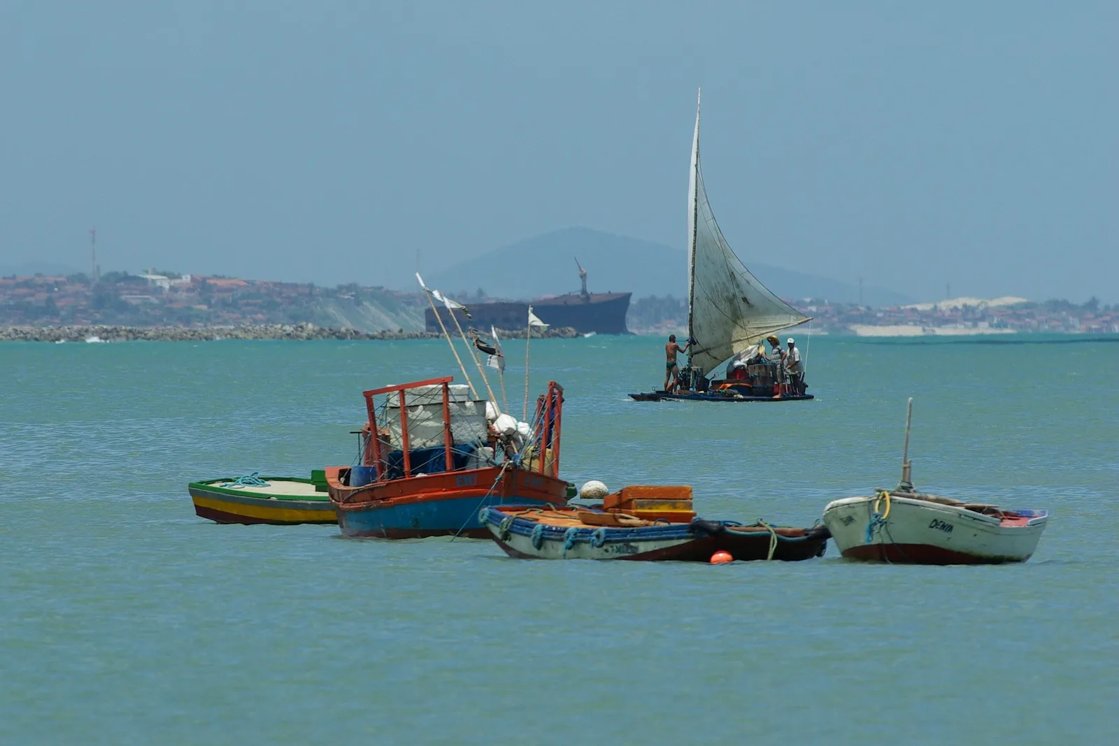 Fishing boats moored in the calm water at Mucuripe with a jangada under a white sail passing behind them, and the cranes and hills of the port on the horizon