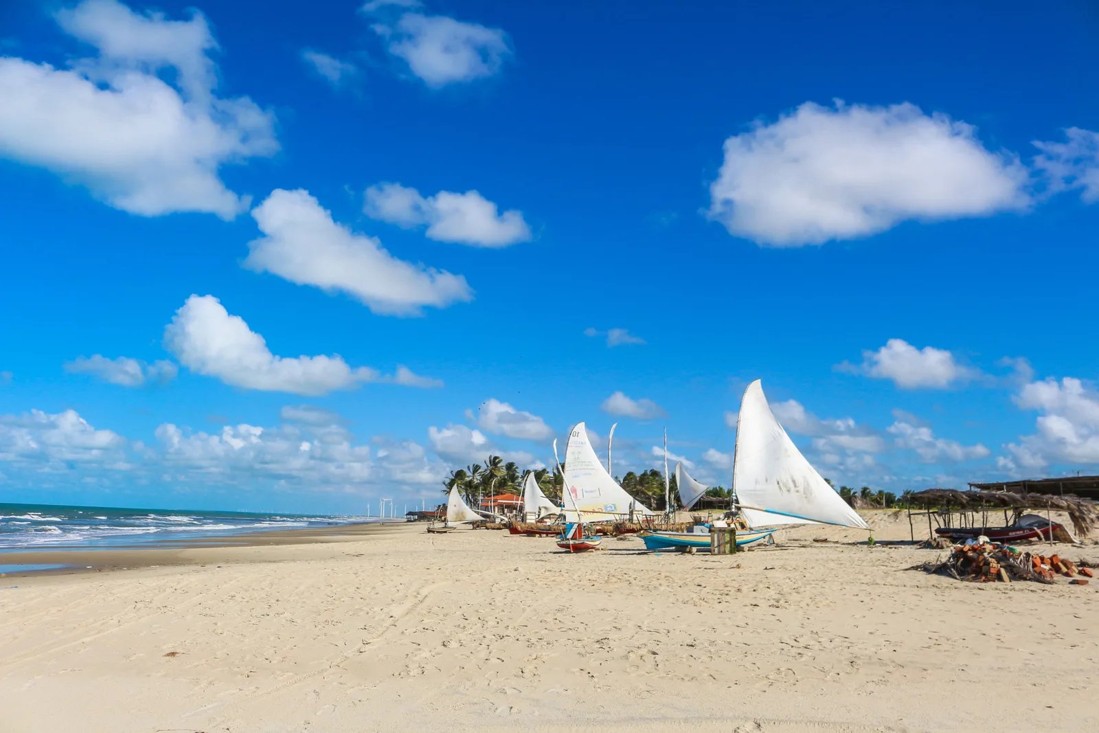 Jangadas with furled white sails and tall masts standing on the pale sand at Praia das Fontes, with flat green sea behind them under a blue sky