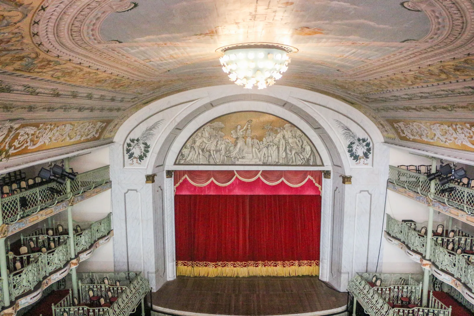 The auditorium of the Theatro José de Alencar: a red curtain under a painted proscenium arch, a chandelier hanging from a decorated ceiling, and tiers of pale cast-iron galleries curving around the room