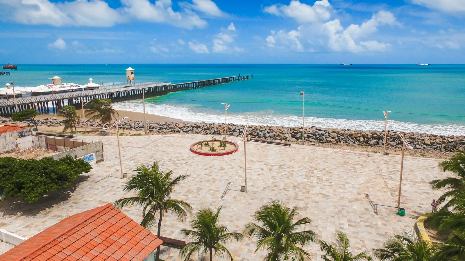A Fortaleza seafront in bright midday sun: a paved plaza in beige stone with a circular planted bed and lampposts on it, a rock revetment running along the shore, a long pier beyond it and coconut palms and a red-tiled roof in the foreground