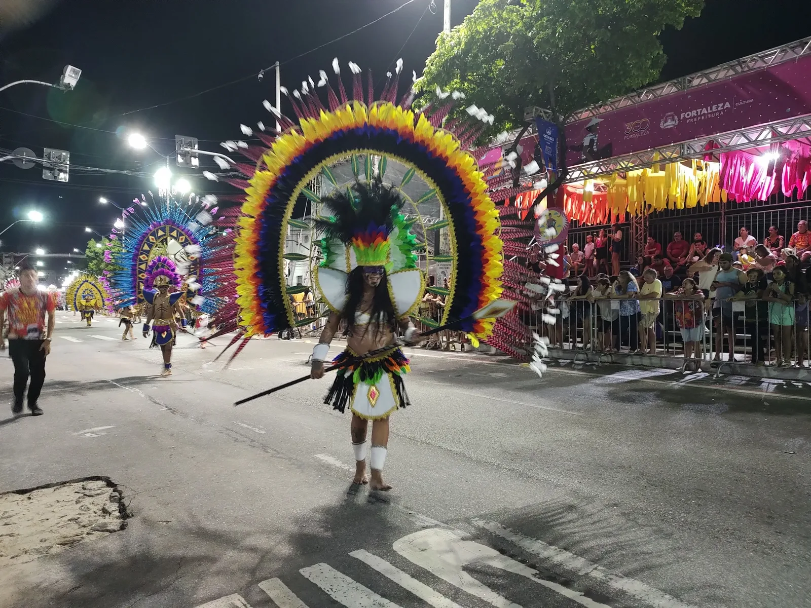 A maracatu performer in a huge fan-shaped headdress of blue, green and yellow feathers walking down a floodlit street at night, with a crowd watching from behind barriers