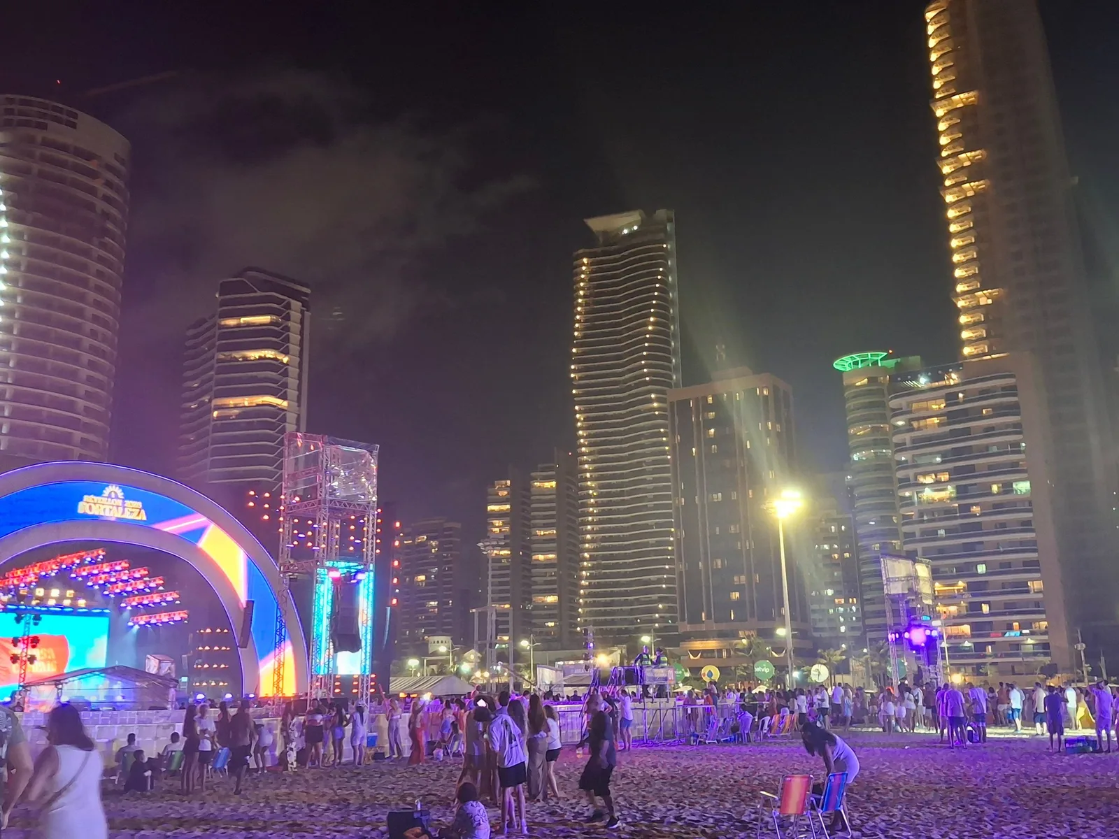 A night crowd on the beach at Praia de Iracema under a lit arch and the floodlit towers of the seafront, with stage lighting throwing purple and green across the sand