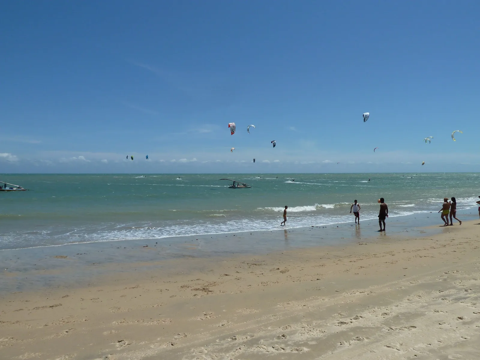 Two dozen kitesurfing kites scattered across the sky over shallow water at Cumbuco, with several people standing ankle-deep on the wet sand in the foreground